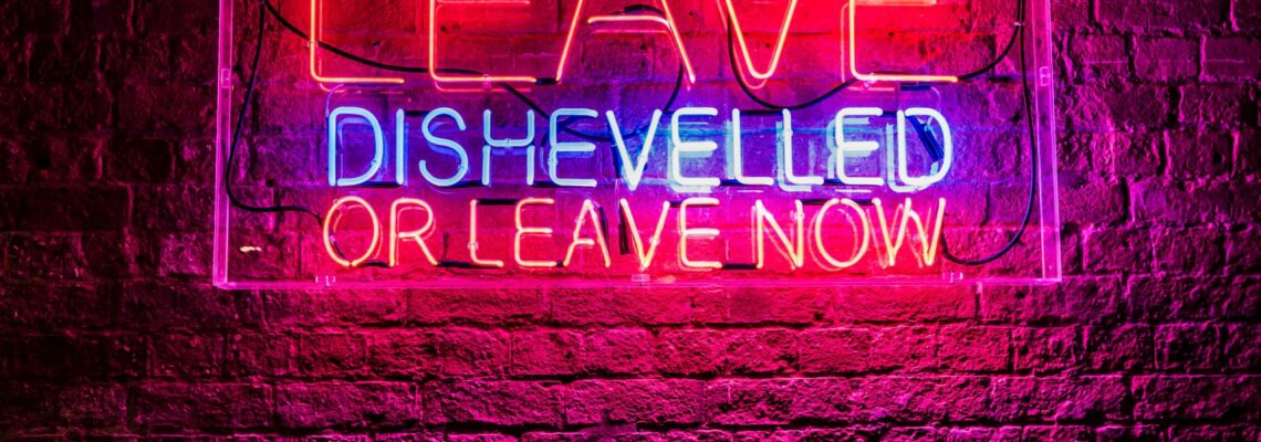 Red and blue neon on a birkc wass reads "Leave dishevelled or leave now."