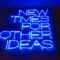 Blue text in neon on a brick wall reads "New times for other ideas." The photo is shot from below.
