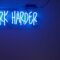 Blue neon off-set at the top right of a blue wall. Text reads, "Work harder."